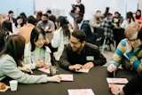 Group of young adults sitting at tables, smiling and playing a game together during an indoor event with many people in the background.
